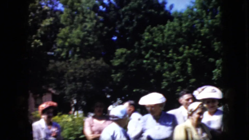 California 1969 8mm frame showing formally dressed adults gathered in bright sun beside greenery, with several women wearing hats and men in suits partly visible.