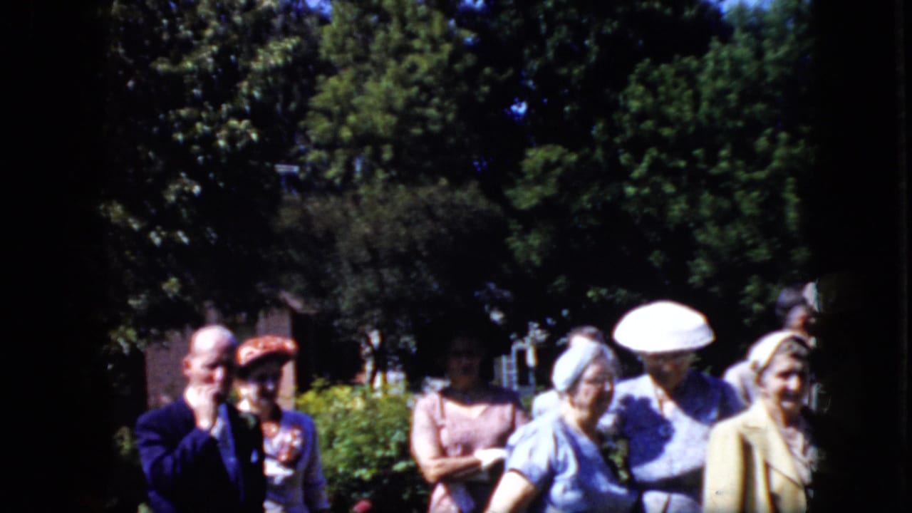 Group of adults in suits, dresses, and hats stands near shrubs under trees.