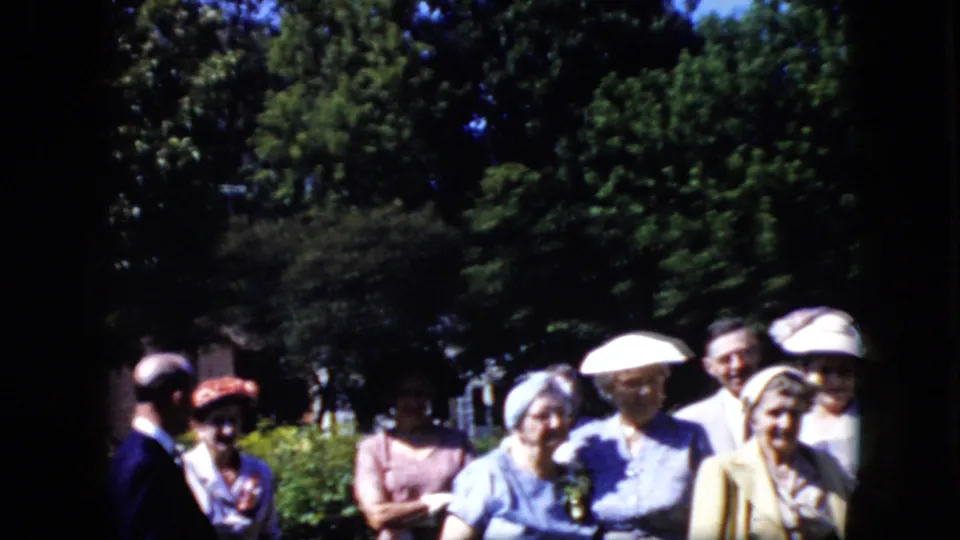 California 1969 8mm frame showing a loose group of formally dressed adults outdoors, with hats, dresses, suits, shrubs, and tall trees visible in soft handheld focus.