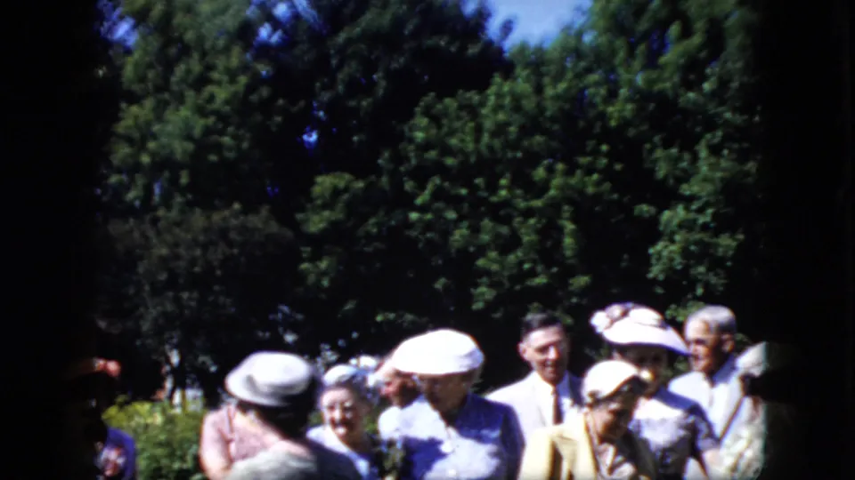 California 1969 home-movie frame showing formally dressed adults clustered near dense green trees and shrubs, with several hats and suits visible in a candid outdoor moment.