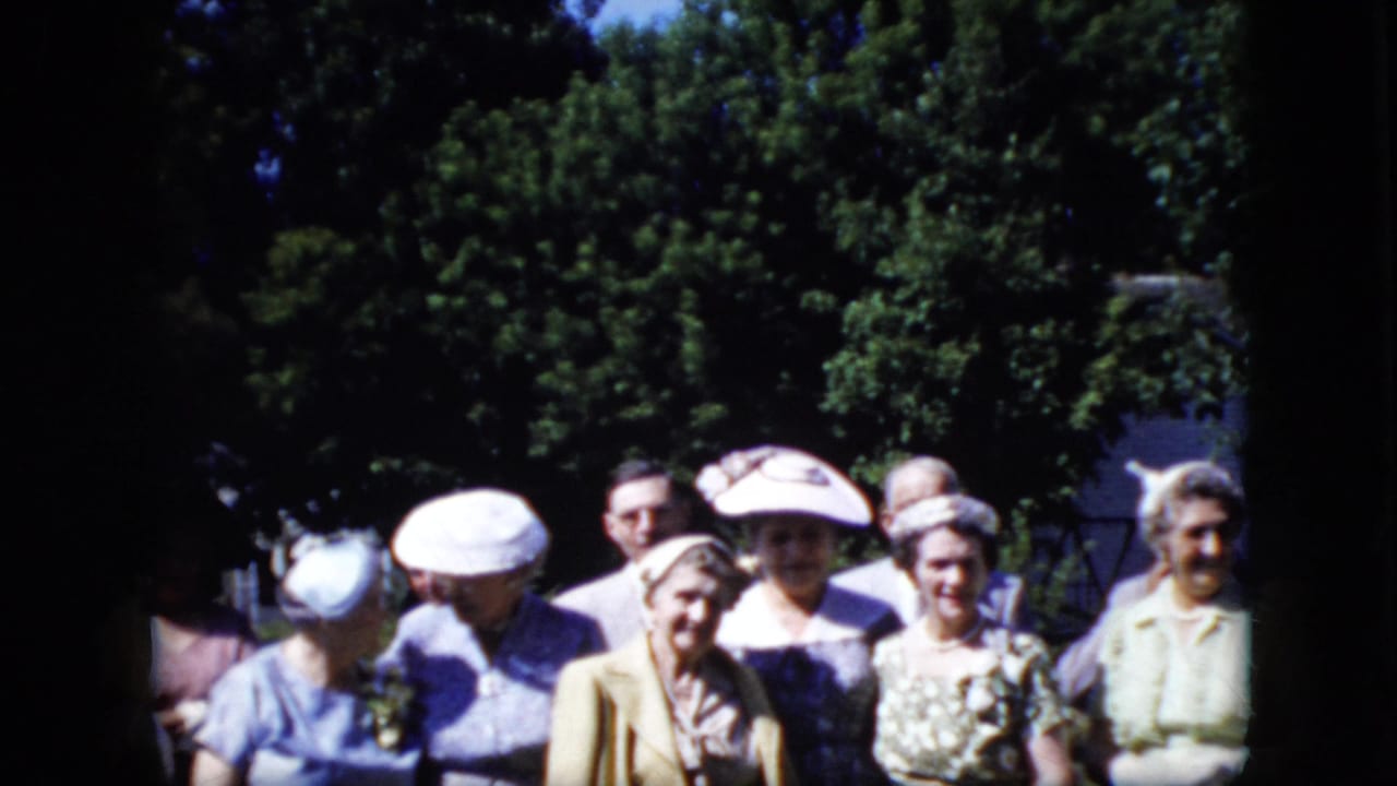 A group of adult women in late-1960s-style hats and dresses face the camera outdoors, with trees and a partial building visible behind them.