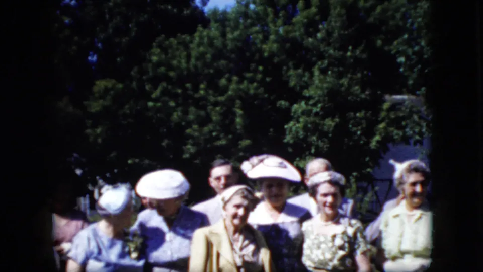 A group of adult women in late-1960s-style hats and dresses face the camera outdoors, with trees and a partial building visible behind them.