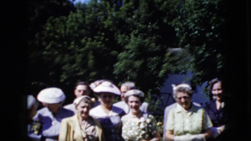 Several adult women in formal clothing stand in a loose row outdoors in 1969 California, including one wearing a floral dress and holding flowers.