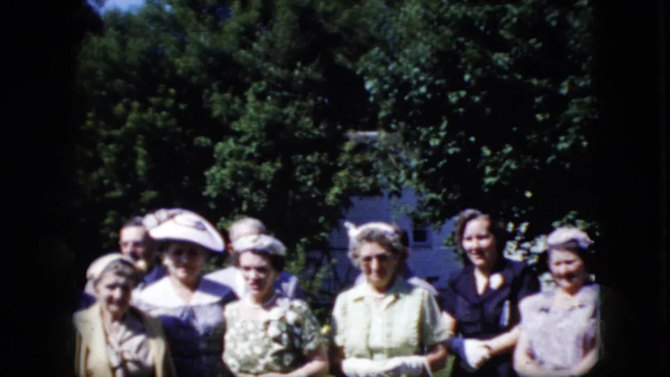 Adult women in dressy late-1960s clothing pose outdoors for the home-movie camera, with hats, floral fabrics, and dense greenery visible around them.