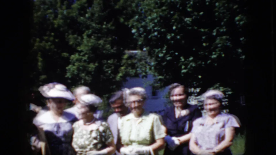 Older adults in formal dresses, hats, and suits appear to pose outdoors in a California garden in 1969, with trees and a white house behind them.
