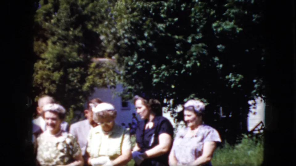 Several older adults in formal clothing are gathered outdoors in a sunlit California garden in 1969, with leafy trees and a white house in the background.