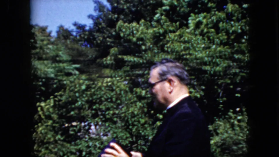 A man in a dark suit and glasses stands in profile in a leafy California garden setting from 1969, holding a small dark item near his hands.