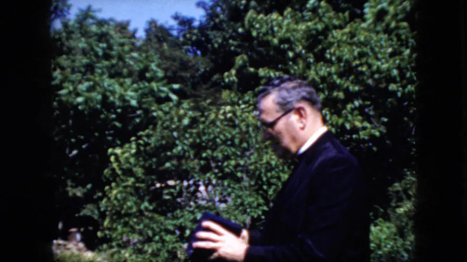 A suited man with glasses appears beside dense garden greenery in 1969 California home-movie footage, holding what may be a book or program.
