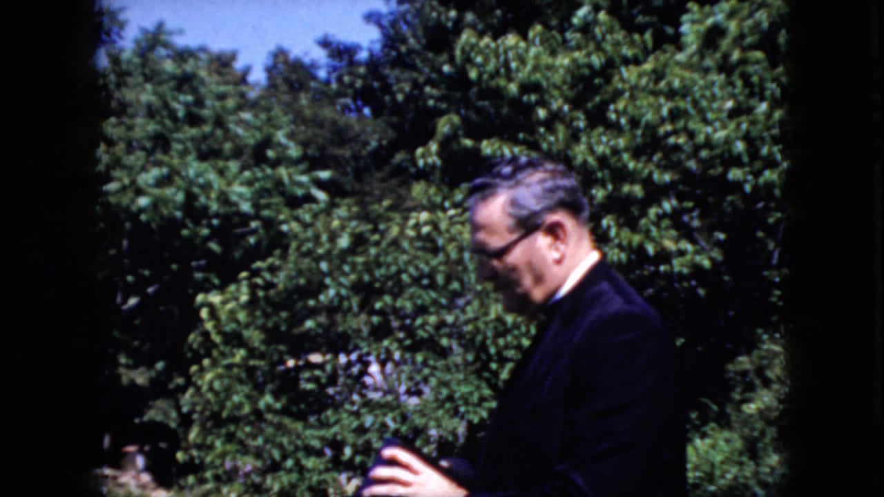 Adult man in a dark suit and glasses stands outdoors in profile against dense green trees in 1969 California home-movie footage.