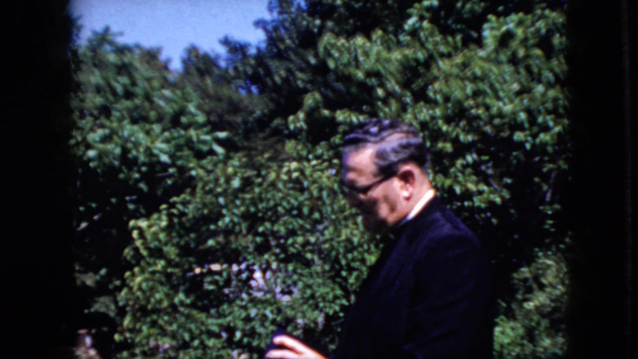Man in glasses looks downward while standing in front of green trees.