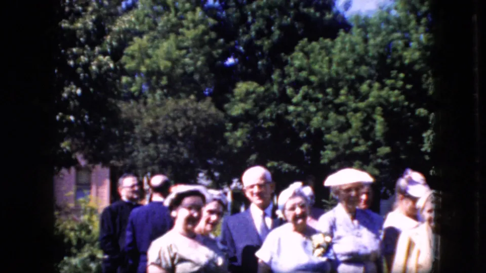 A group of adult men and women in late-1960s-style suits, dresses, and hats stands outdoors among trees in the supplied California 1969 footage.