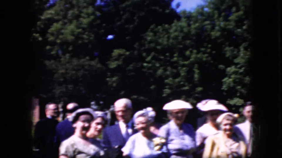 The home-movie frame shows adults gathered in a leafy outdoor setting, with women in hats and dresses and men in suits visible in soft 8mm color.
