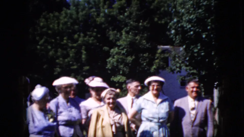 Several adult attendees, including women in light hats and men in suits, stand together in a bright outdoor garden or park setting in the supplied 1969 California clip.