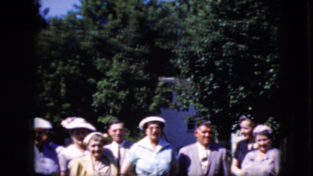 Older adults standing outdoors in bright sun and shade with trees and a white building behind them.