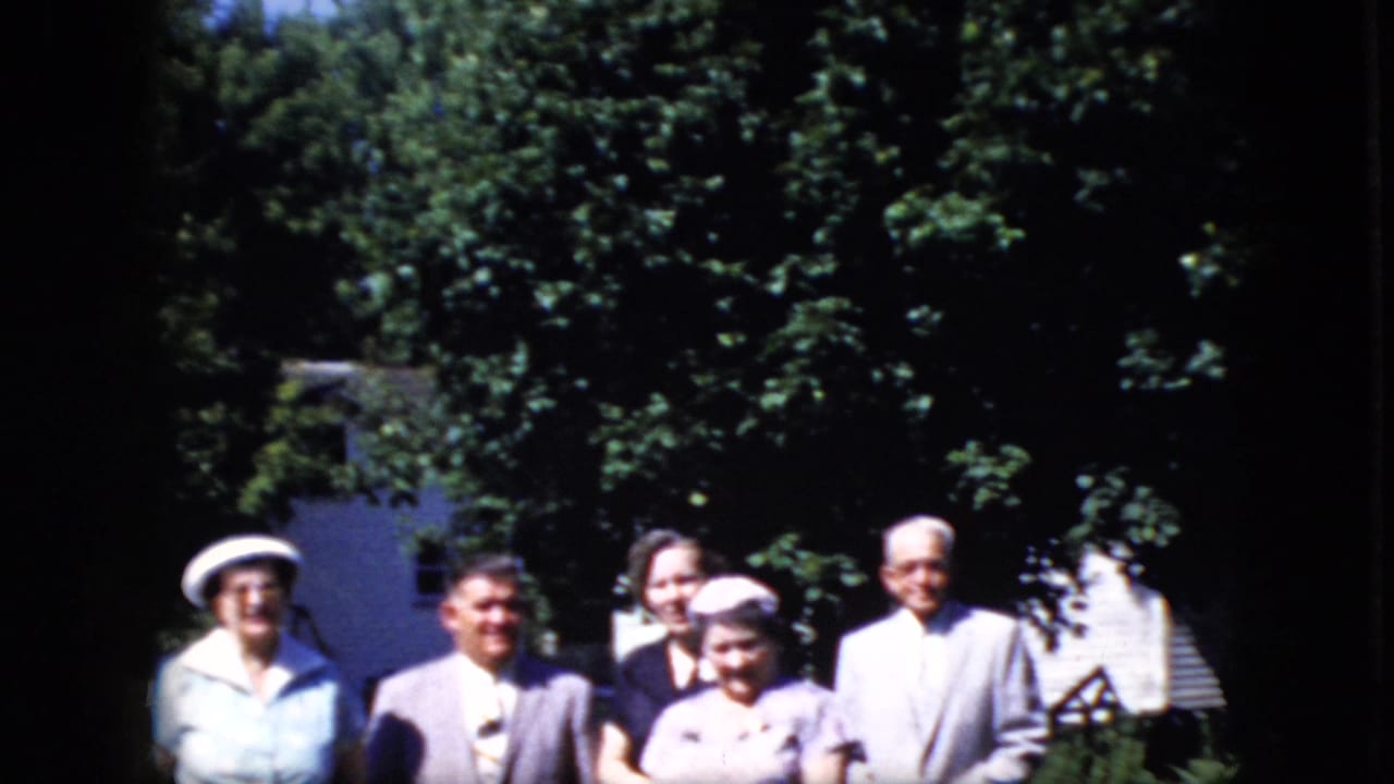 Smaller group of older adults posing under trees with a white building partly visible behind them.