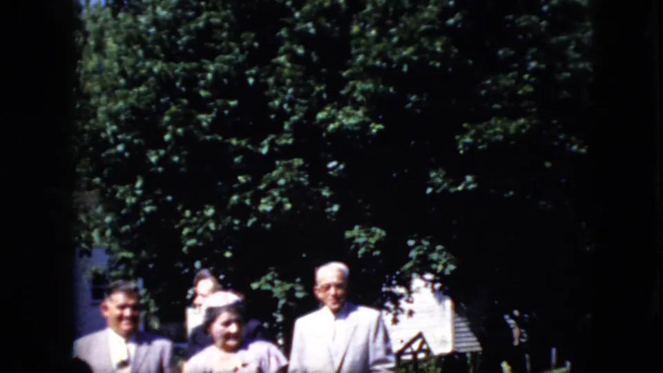 1969 California 8mm frame with several older adults partly cropped at the lower edge, standing outdoors below dense trees beside a white building.