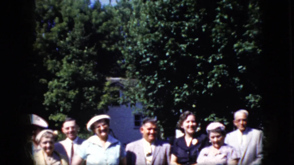 1969 California home-movie frame showing older adults in period dresses, hats, and suits standing together outdoors near trees and a white building.