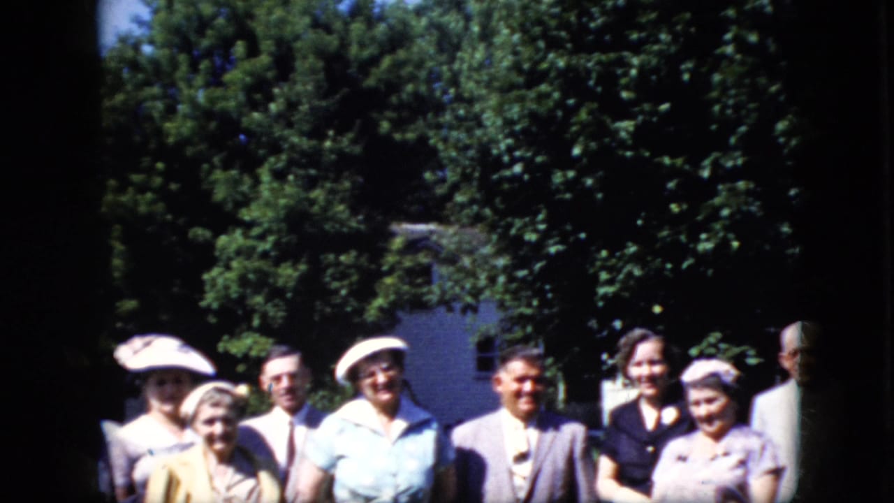 Adults standing outdoors in front of trees and a house, with several women in hats and men in suits.