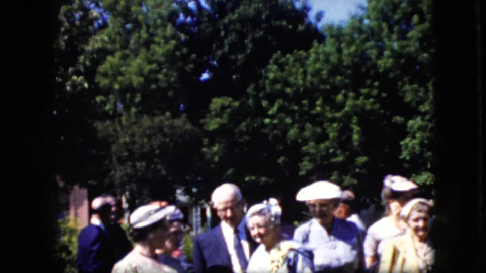 Older and mature adults in California in 1969 stand together in a shaded yard, with a suited man, women in hats, leafy trees, and residential surroundings visible.