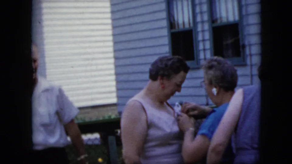 Two mature women in a California residential yard in 1969 stand close together near the white-sided house, with other adults partially visible at the frame edges.