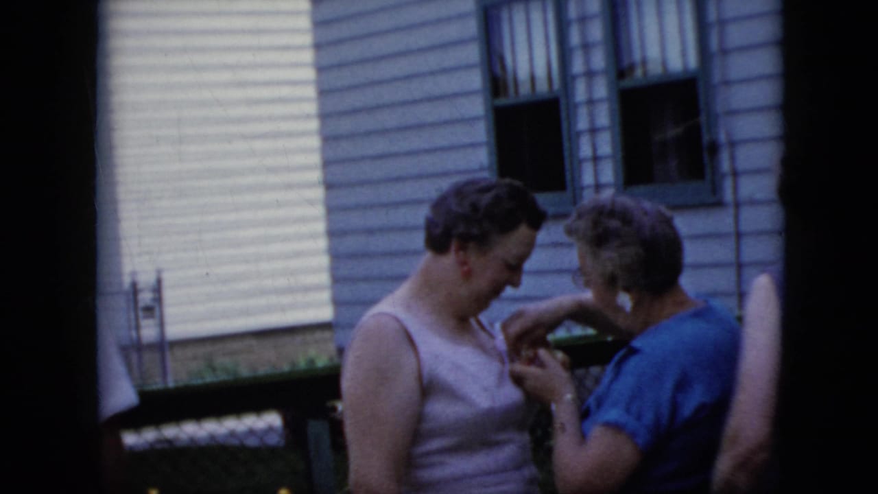 In 1969 California home-movie footage, two adults stand near a house exterior while one appears to adjust a corsage on the other's sleeveless dress.