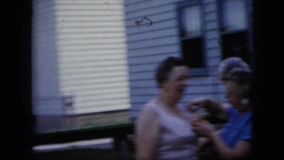 Two adults remain close together outside a California residence, continuing the corsage adjustment in soft, handheld 8mm footage.