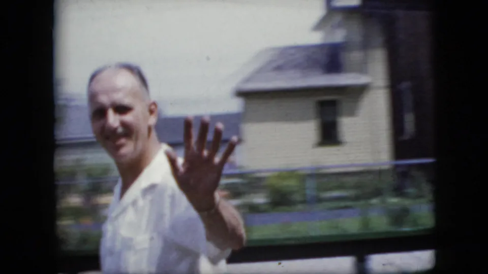 A man in a white shirt smiles and waves toward the home-movie camera in a California residential setting, with houses and yard fencing behind him.