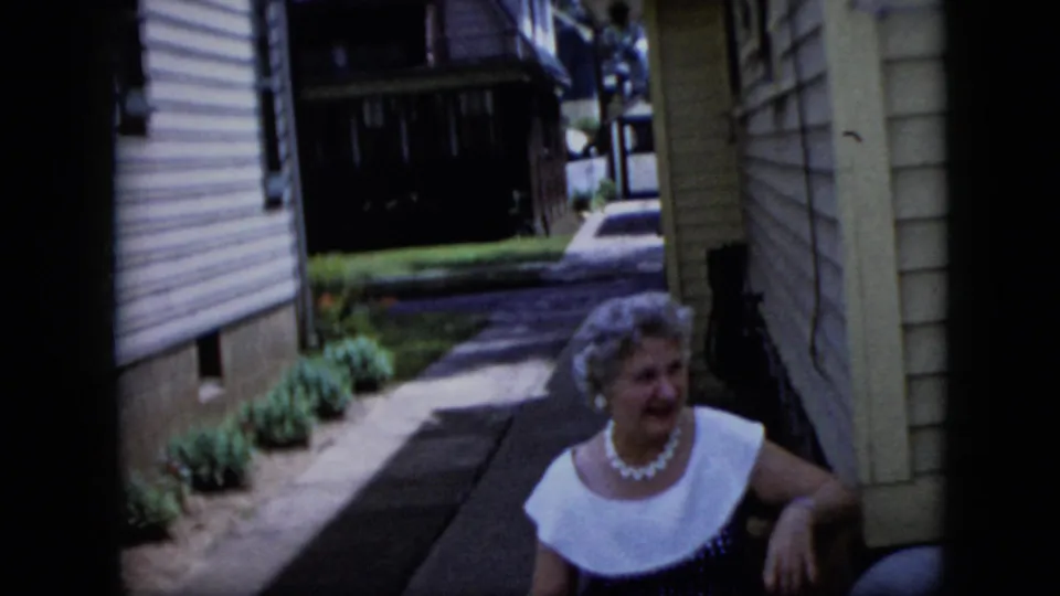 An older woman in a white-collared dress sits near a porch or doorway in a California residential walkway scene recorded on 8mm film.