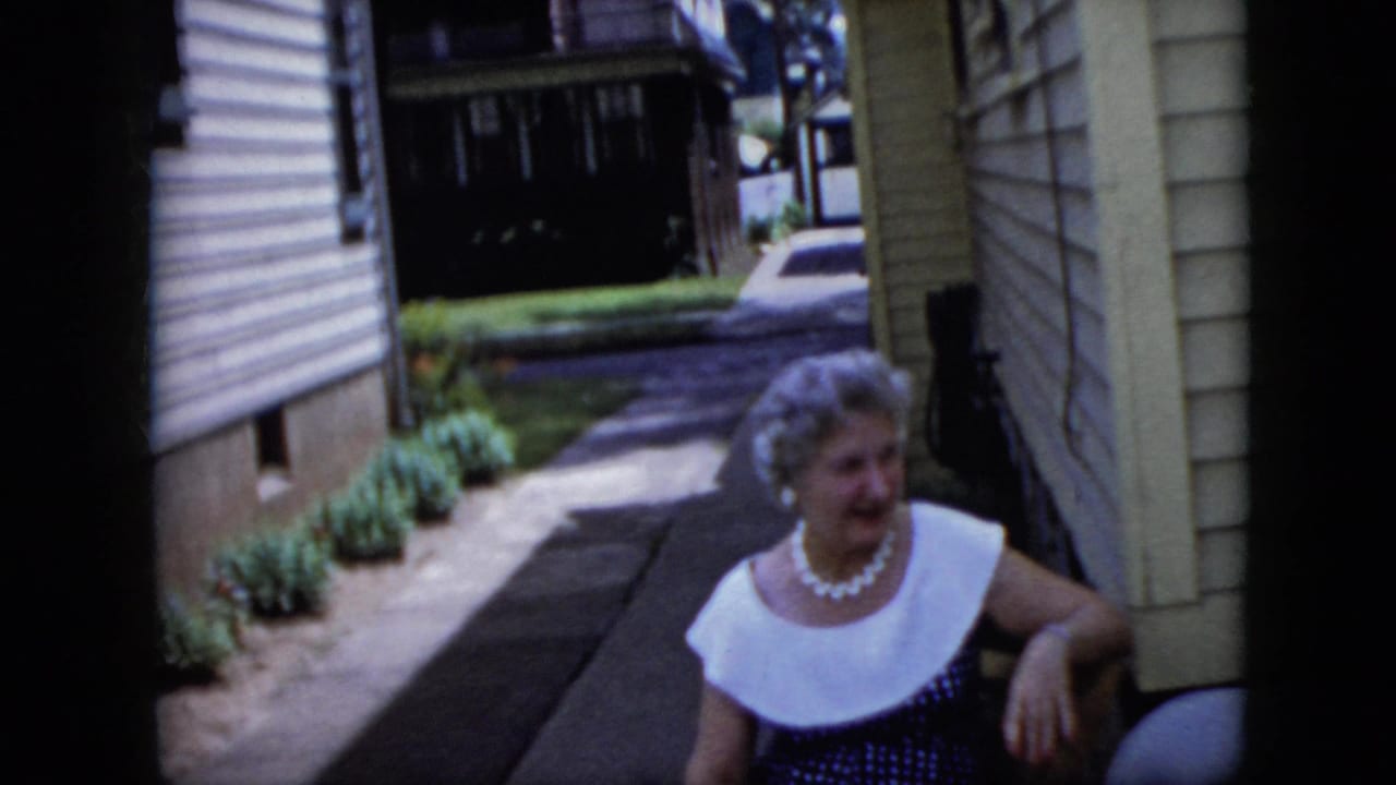 Grey-haired woman in a pearl necklace seated beside a narrow residential walkway.