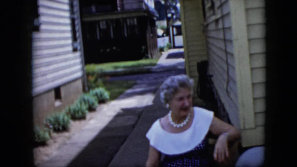 In 1969 California home-movie footage, a grey-haired woman wearing pearls and a white collar sits near house siding beside a narrow concrete walkway.