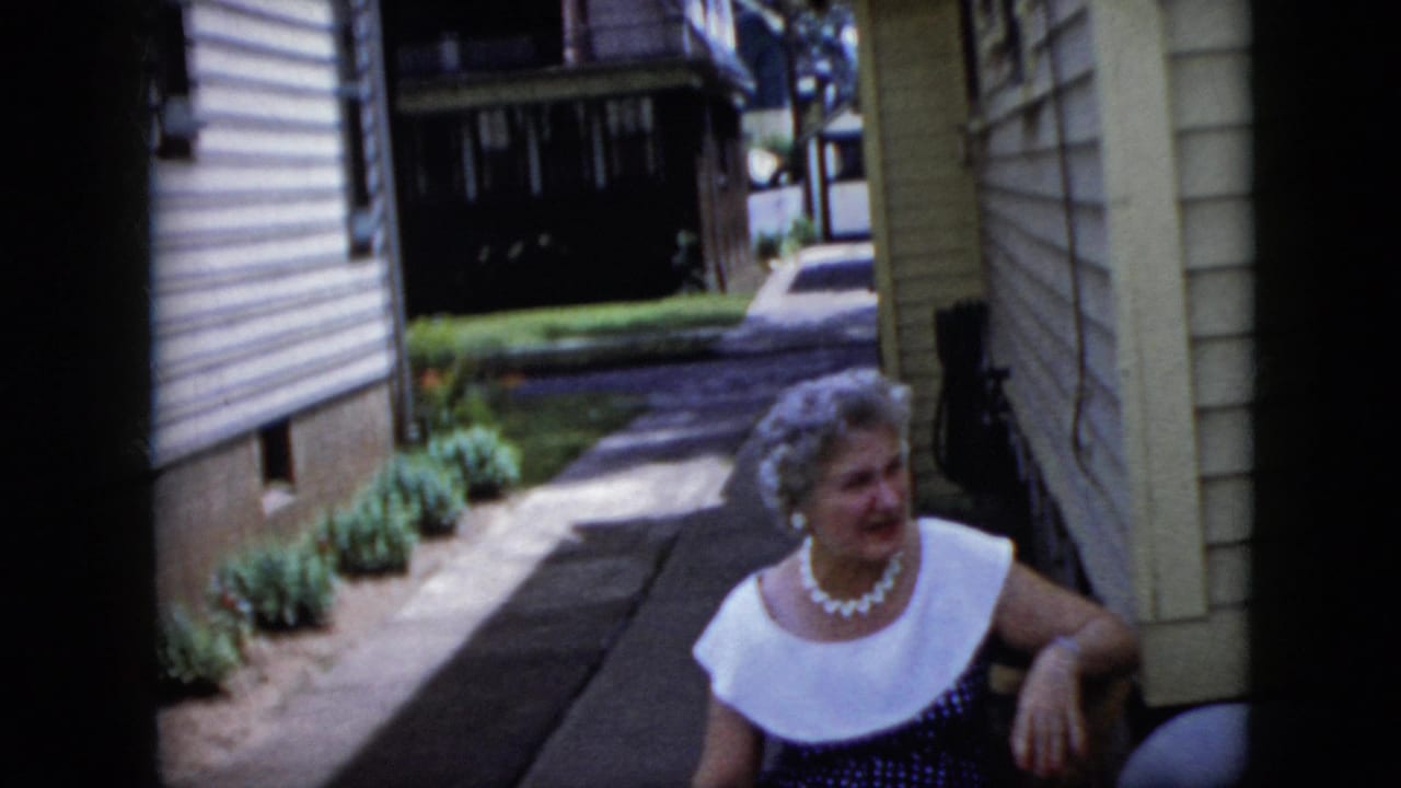 Seated woman turns toward someone off-camera in a narrow passage between houses.