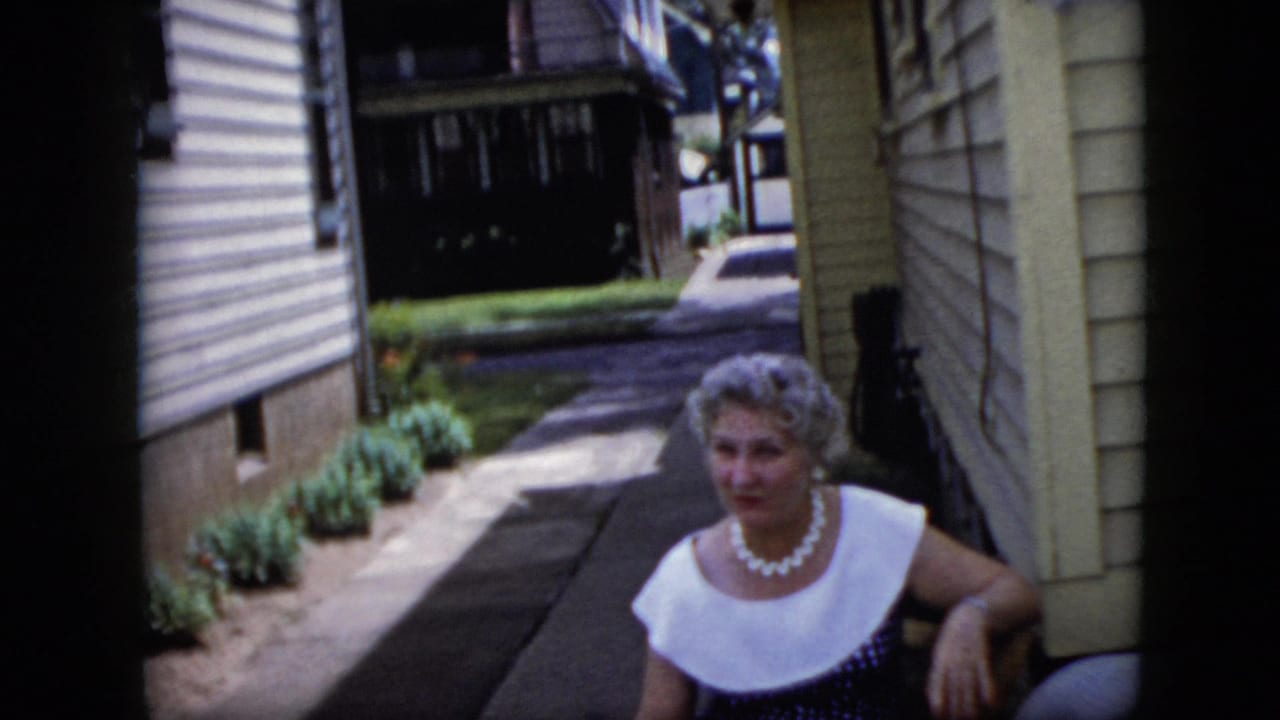 Older woman in a white-collared dress sits near a house walkway.