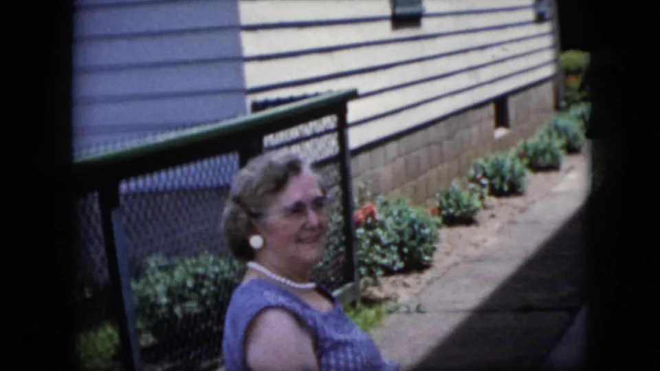 The woman in glasses and a blue sleeveless top is seen closer to the camera beside the chain-link fence, with residential siding, plantings, and a concrete path behind her.