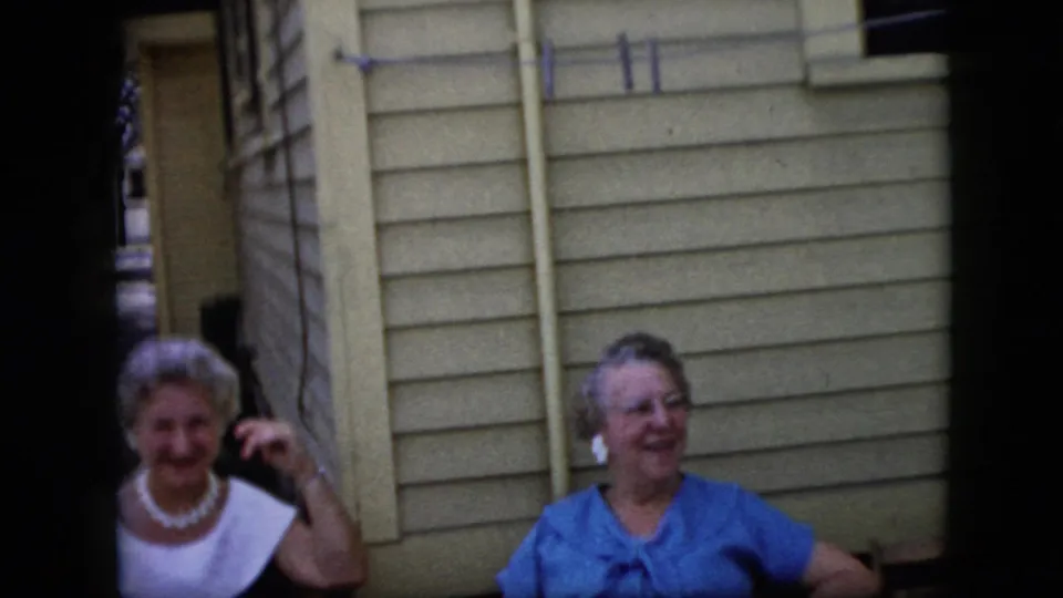 Two older women sit outdoors near light-colored house siding, one in a white-collared dress and one in a blue top, both visible in the final home-movie frame.