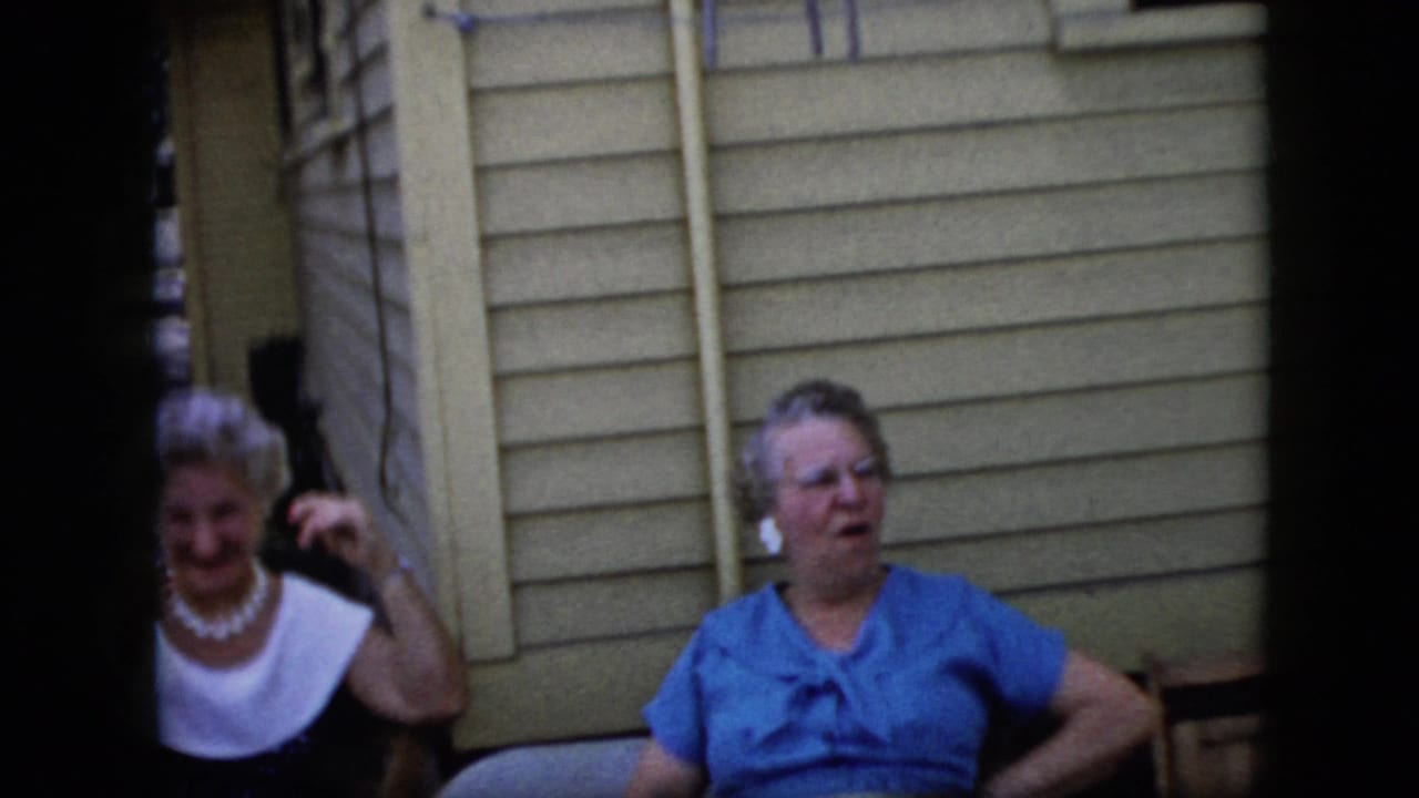 Two older women seated outside a light-sided house, one in a white-collared top and one in a blue blouse.