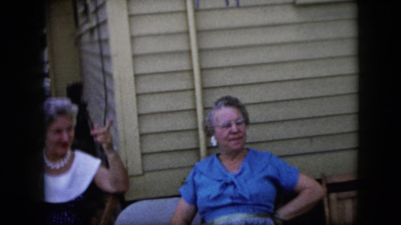 Two older women sit outside a house while one raises her hand near her face.