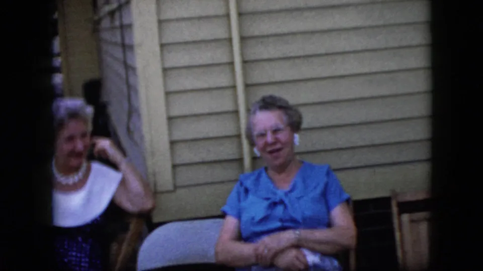Two older women remain seated outside the light-sided house in 1969 California footage, with the woman in blue smiling and folding her hands near her lap.