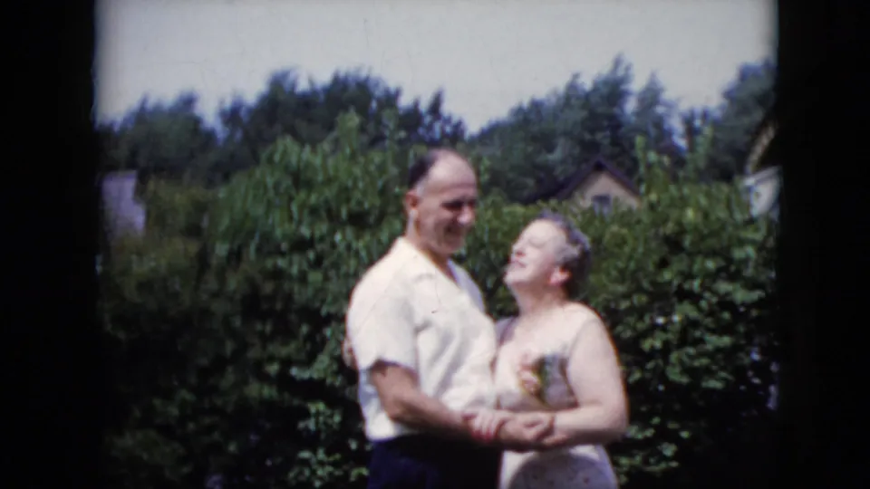 A man and woman stand close together in a California garden in 1969 home-movie footage, embracing in front of dense greenery and a distant house roofline.