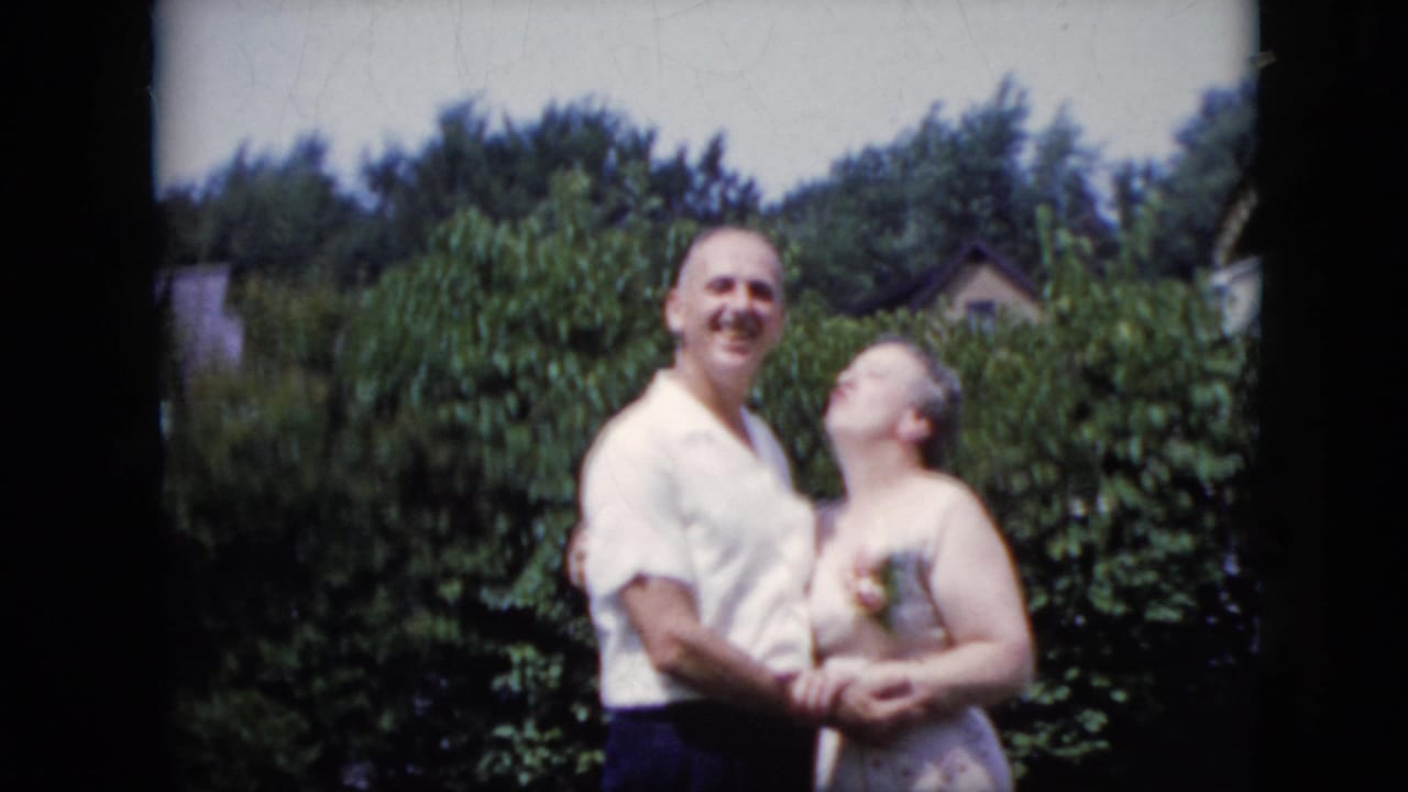 Adult couple standing close together in a backyard with bushes behind them.