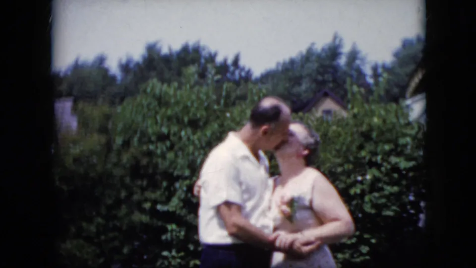 In a 1969 California backyard scene, the adult couple shares a kiss beside leafy bushes while standing close together in daylight.