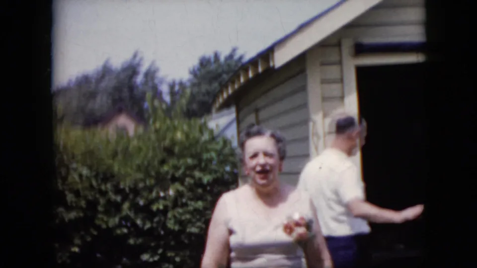 The woman faces the home-movie camera with a corsage visible as the man turns away beside a light-colored backyard building or house wall.