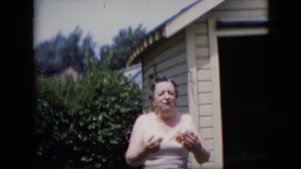 The woman stands alone near leafy bushes and a light-colored residential structure, holding or adjusting the corsage as she faces the camera.