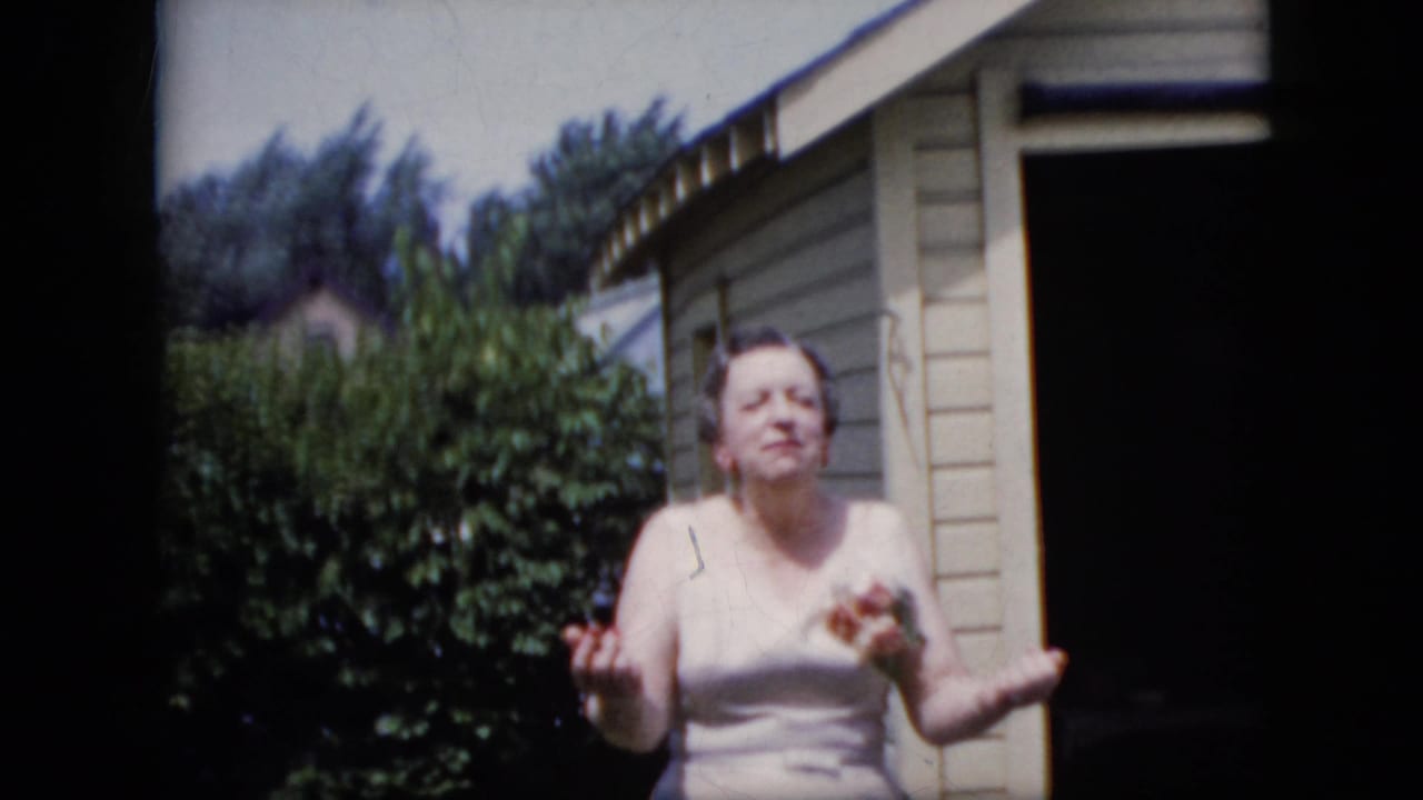In supplied 1969 California home-movie footage, an adult woman in a light sleeveless dress gestures while standing outside a beige-sided residential structure with shrubs behind her.