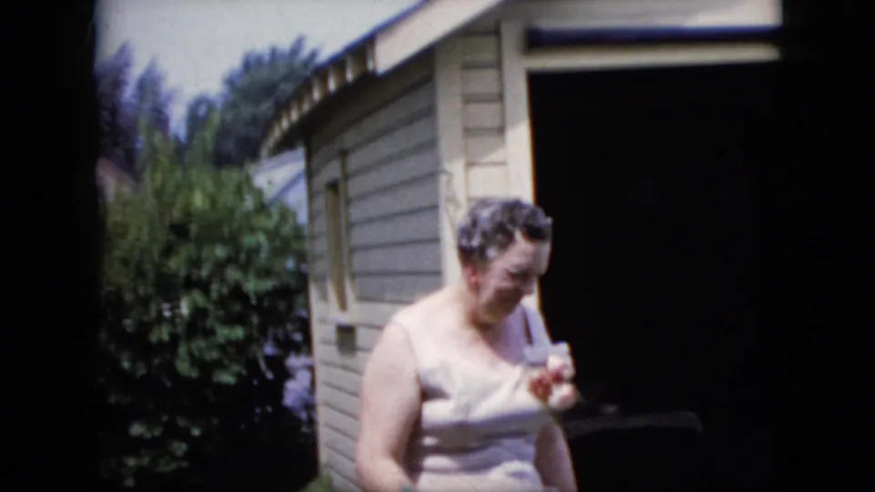 The woman stands beside the beige-sided structure and looks down at a small object in her hand, with the doorway and sunlit shrubs visible around her.