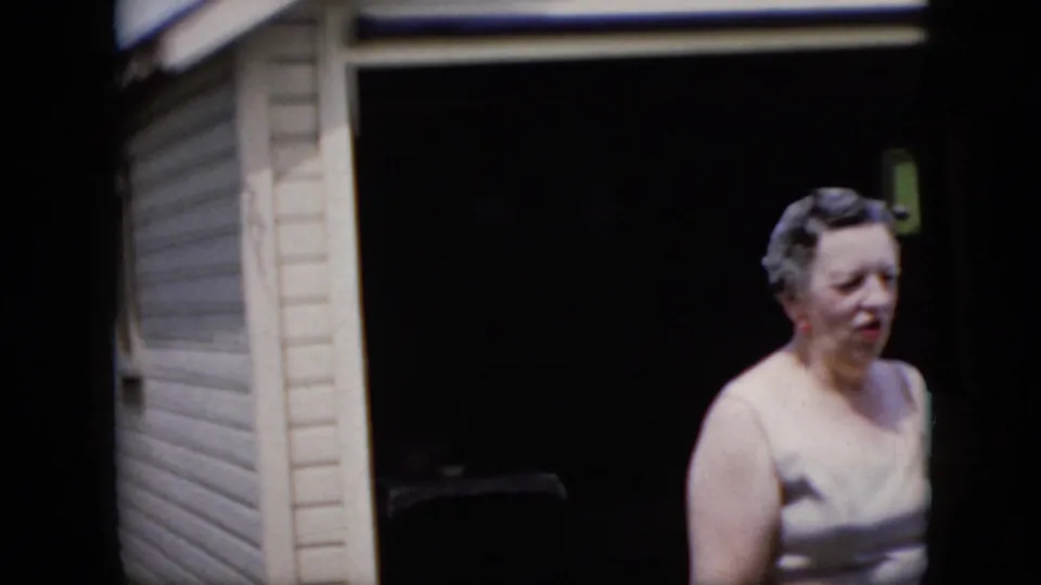 The woman appears in side view near the dark doorway of the beige-sided residential exterior, continuing the candid outdoor conversation recorded on 8mm film.