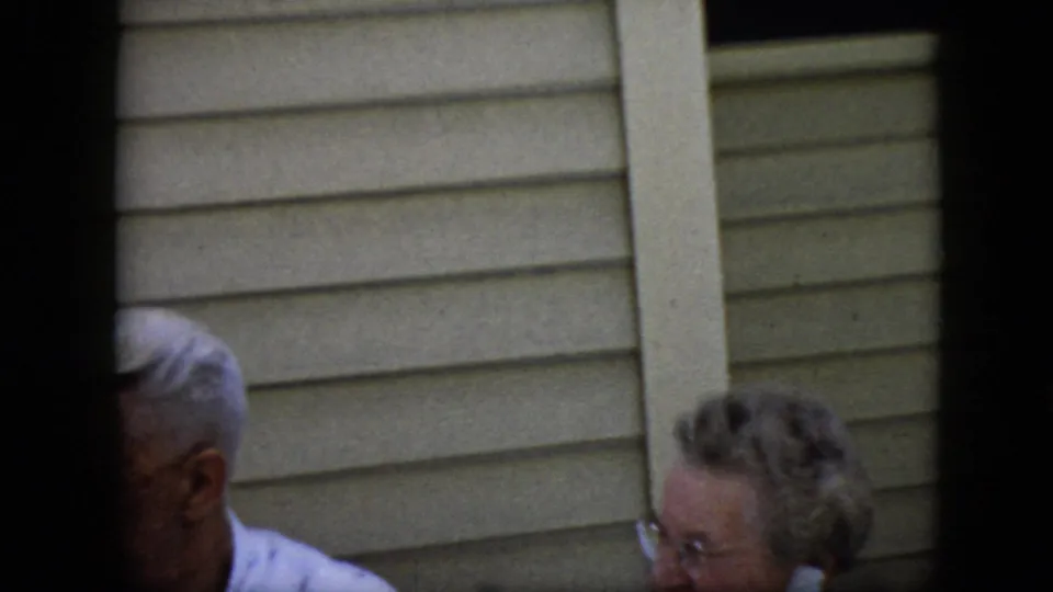 The frame shifts close to the beige siding and catches partial figures near the bottom edge, including an older adult with glasses and another gray-haired person partly obscured.