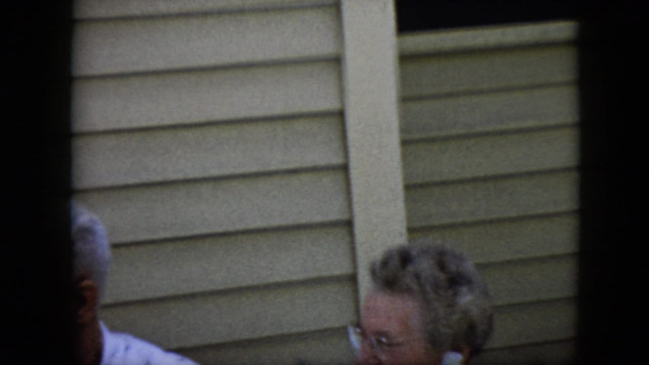 Partial view of an elderly couple near beige house siding, partly obscured by dark film borders.