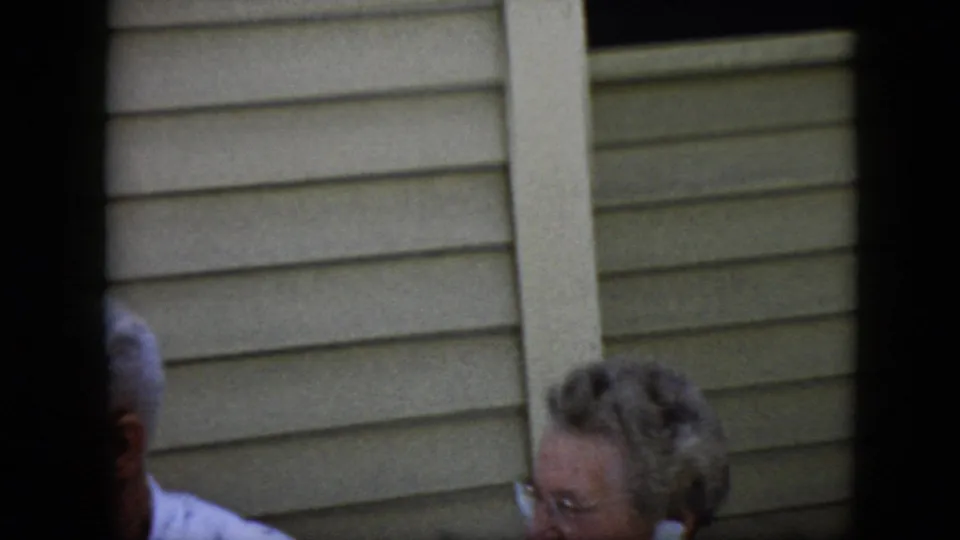 1969 California home-movie frame showing an elderly man and woman partly cut off near beige horizontal siding, with dark 8mm frame edges visible.