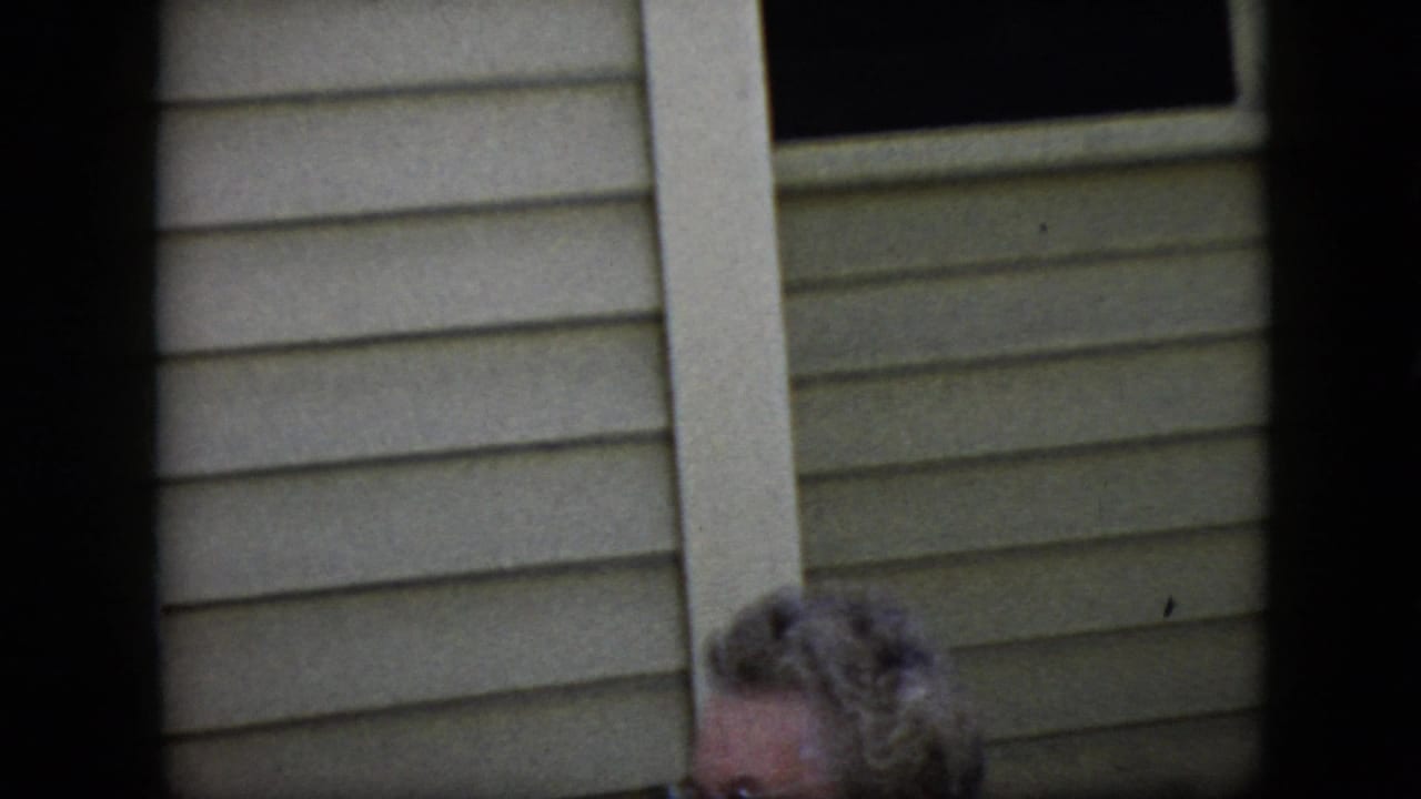 Elderly woman with glasses near horizontal siding, partly hidden by dark frame edges.