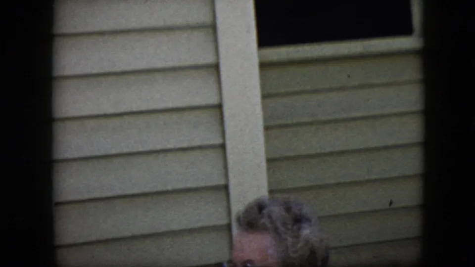 1969 California home-movie frame with an elderly woman in glasses partly visible beside beige siding, her face turned toward something off-camera.